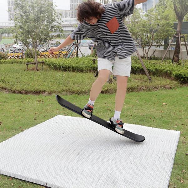 A person on a ski board performing a trick on ski mat in an outdoor setting with greenery and a building in the background.
