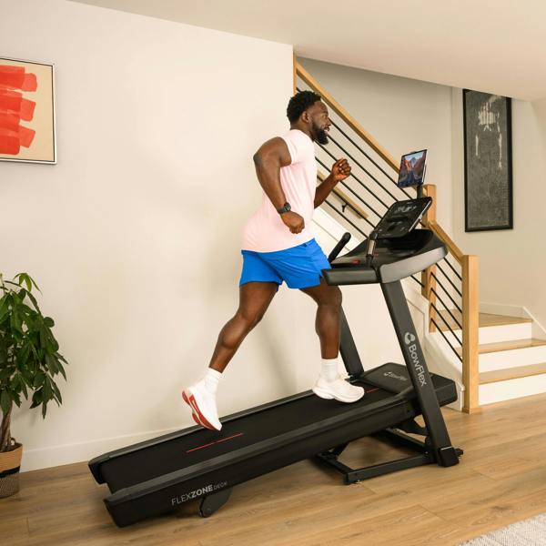 Man running on a treadmill in a home setting with a staircase and plant in the background.