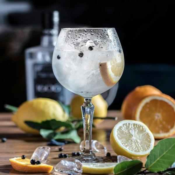 Chilled gin and tonic in a copa balloon glass garnished with lemon, ice, and juniper berries on a wooden table.