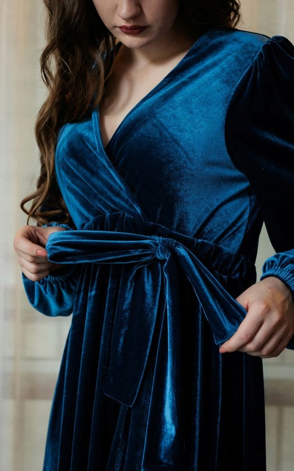 Here is a upper torso close up of the teal velvet dress. The model in the image has brown hair and her hands are holding the two ends of the bow to show that it is adjustable. The gathers and the elasticised cuffs are also exhibited. The background is a cream white. 