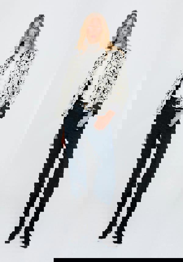 Woman wearing a patterned jacket, white turtleneck, dark jeans, and black boots on a white background