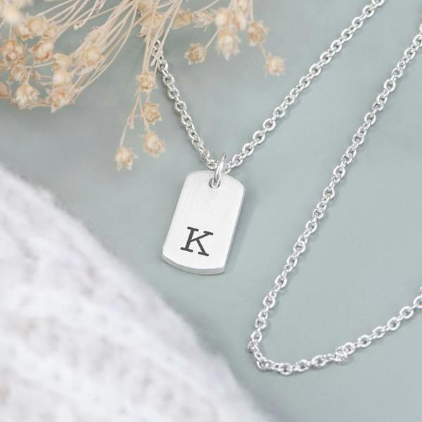 A dainty silver-plated rectangular tag pendant necklace engraved with a custom initial letter, laid flat on a clean surface.