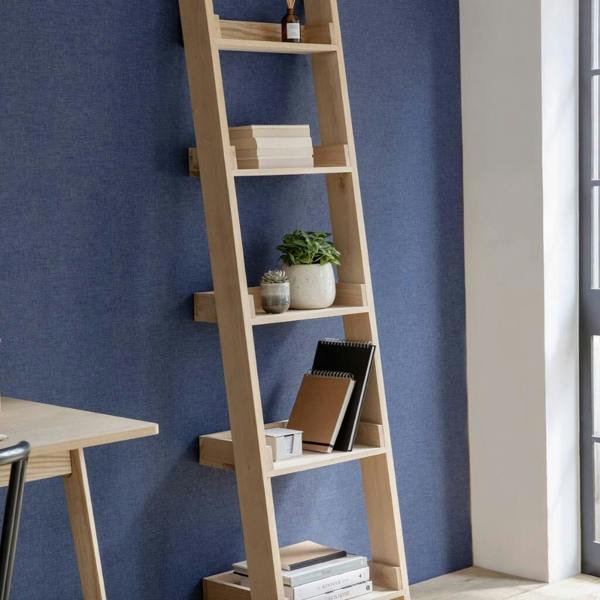 A minimalist raw oak shelf ladder unit stacked with books, plants, and notebooks against a textured dark blue wall.