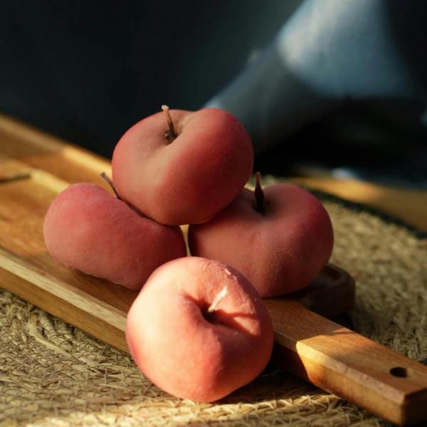 Four unlit, realistic flat peach-shaped pink candles with a fuzzy, velvety texture stacked on a wooden serving paddle in dramatic, sunlit studio lighting.