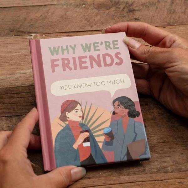 A person holding the 'Why We’re Friends' hardback gift book. The pink cover features an illustration of two friends sharing coffee with a speech bubble that reads '...you know too much.' The book is set against a rustic wooden background.