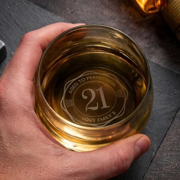 Hand holding a glass whiskey tumbler filled with amber spirit, showing a personalised circular engraving on the base that reads 'Aged to Perfection 21' with a custom message.