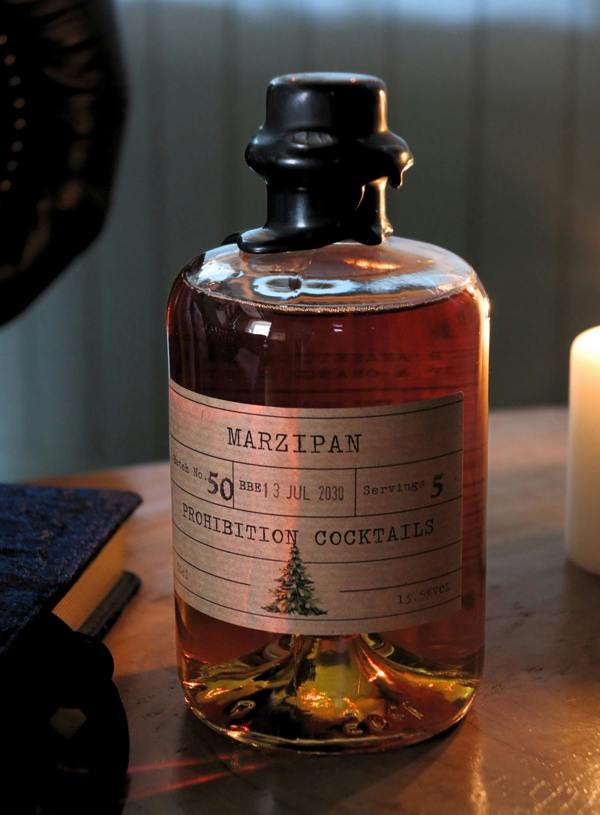 A clear glass bottle filled with amber liquid and topped with a black cap, standing on a wooden surface. The bottle has a cream label reading 'MARZIPAN' and 'PROHIBITION COCKTAILS' with a small tree illustration.