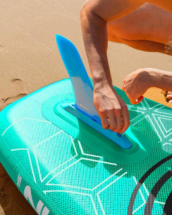 Person attaching a blue fin to a green inflatable paddleboard on sandy beach