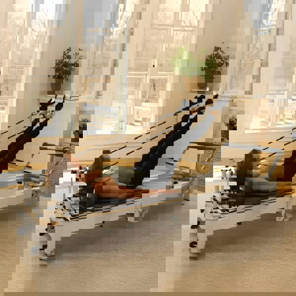 Aluminium Pilates reformer machine with a padded platform, resistance cords, and a person performing leg exercises in a bright room