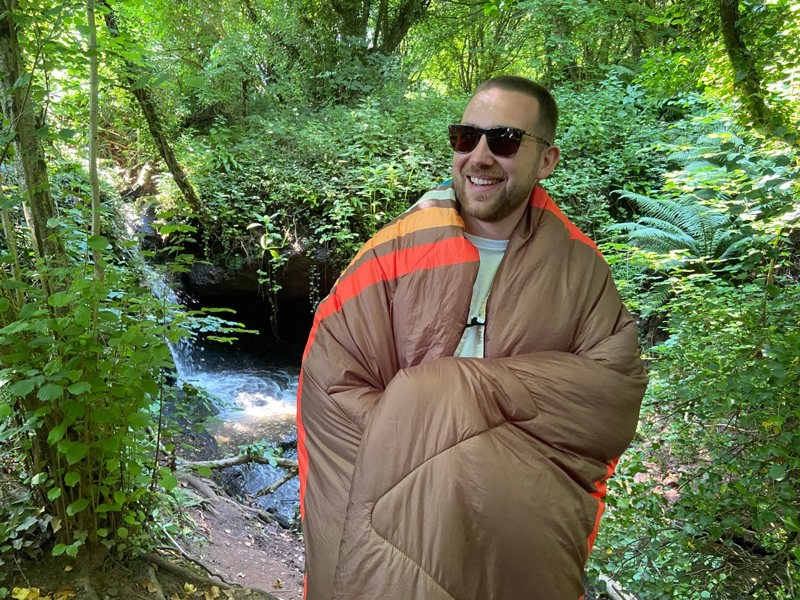 Vivid spruce Design Blanko being worn by a young man with the outdoor blanket wrapped around him walking in the country side. imageset:Vivid Spruce
