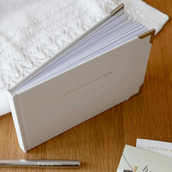 Personalised Christening Guest Book standing upright on a wooden table, silver-foiled with BABY OLLIE and displaying its thick paper pages.