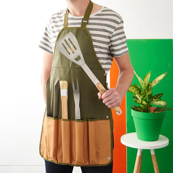 A man wearing an olive green canvas barbecue apron holding a large stainless steel spatula, with tongs and brushes stored in the front pockets.