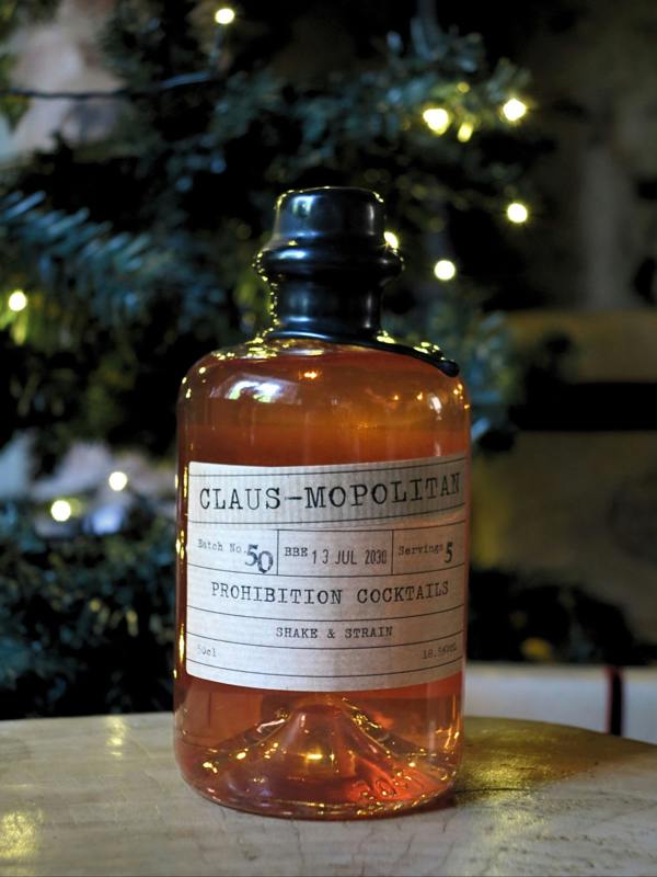 A vintage-style amber glass bottle with a black cap and a white label reading 'CLAUS-MOPOLITAN PROHIBITION COCKTAILS', photographed on a tabletop with blurred Christmas tree lights in the background.