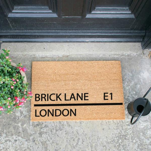 A natural coir street sign doormat reading Brick Lane E1 London outside a black front door.