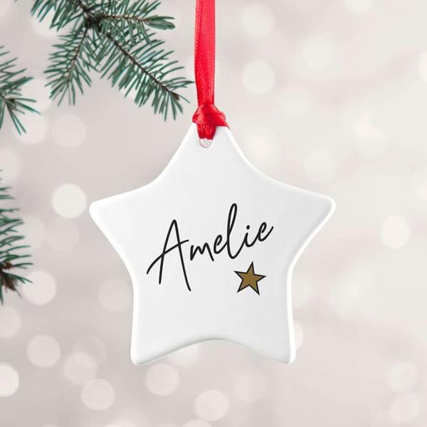 A personalised white ceramic star Christmas tree decoration with a custom name and a small gold star, hanging from a red ribbon.