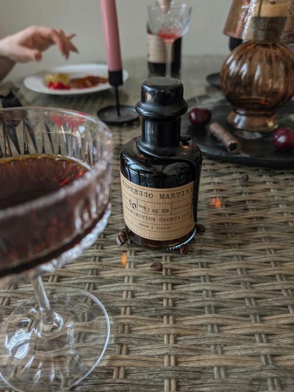 A small dark glass bottle labeled 'ESPRESSO MARTINI' sits on a woven table mat beside a stemmed glass filled with dark red liquid, with candles, cherries, and other bar-like props in a softly blurred indoor setting.