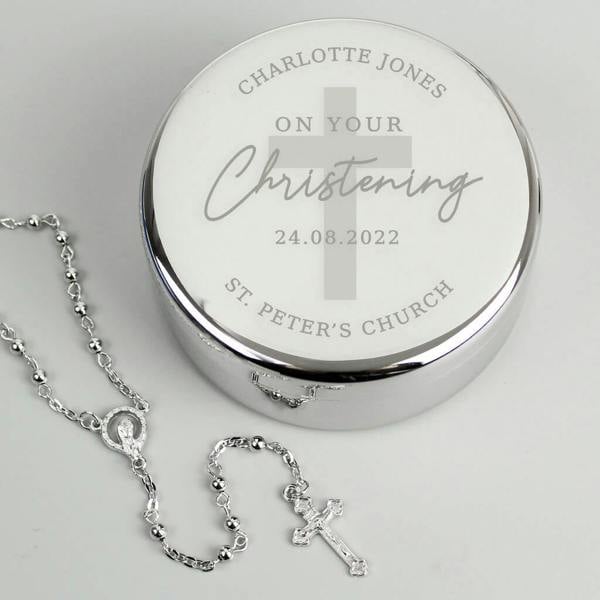 A silver, circular personalised Christening trinket box engraved with "Charlotte Jones," "On Your Christening," a date, and "St. Peter's Church," with a delicate silver rosary bead set laid beside it on a neutral background.
