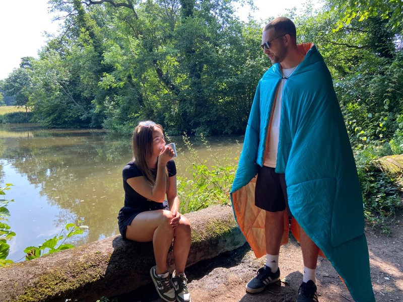 A young couple in the outdoors with one of them stood with an insulated reversible Blue/Orange Blanko outdoor blanket.