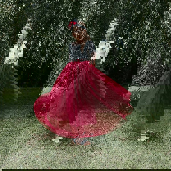 Young girl in a white top and red skirt standing outdoors with greenery in the background
