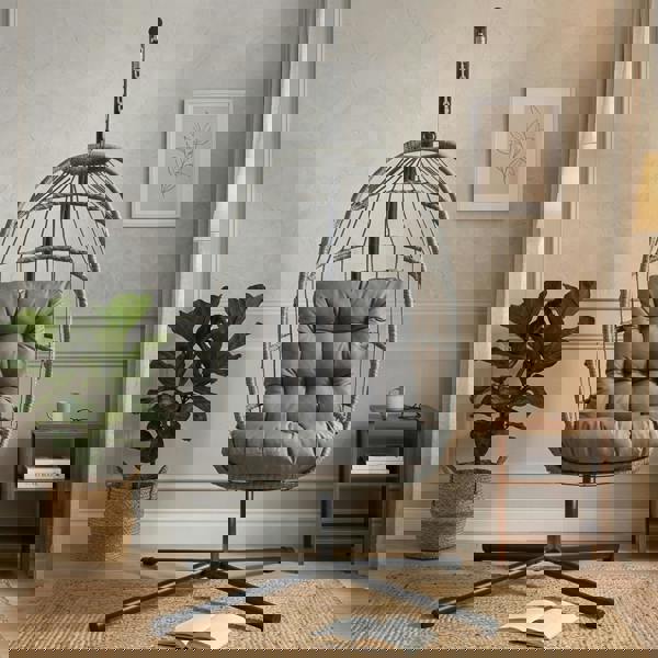 Grey rattan hanging egg chair with plush cushion, suspended from a sleek metal stand, set in a cozy, modern living room environment