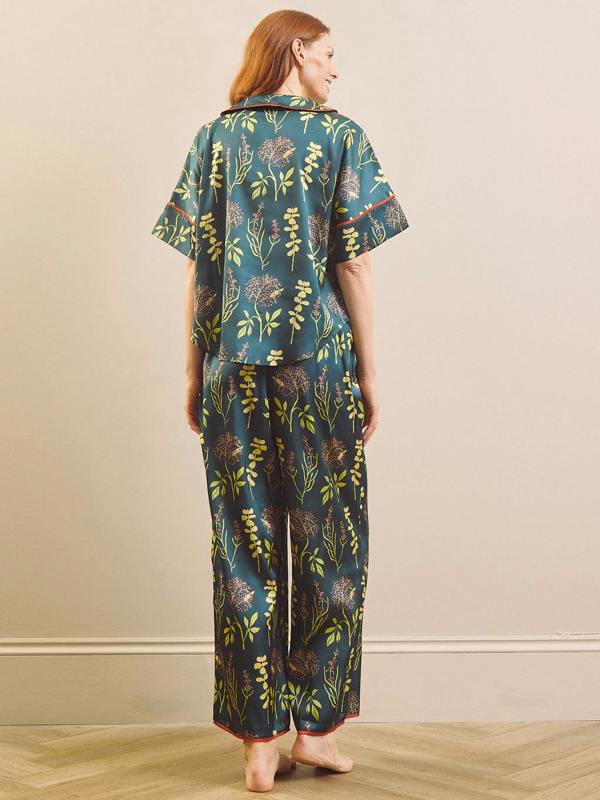 A woman stands barefoot indoors wearing a matching short-sleeve top and wide-leg pants pyjama set in a dark teal green color with a yellow and brown botanical pattern.