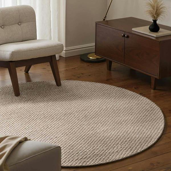 Round beige rug in a living room with a chair and wooden cabinet.