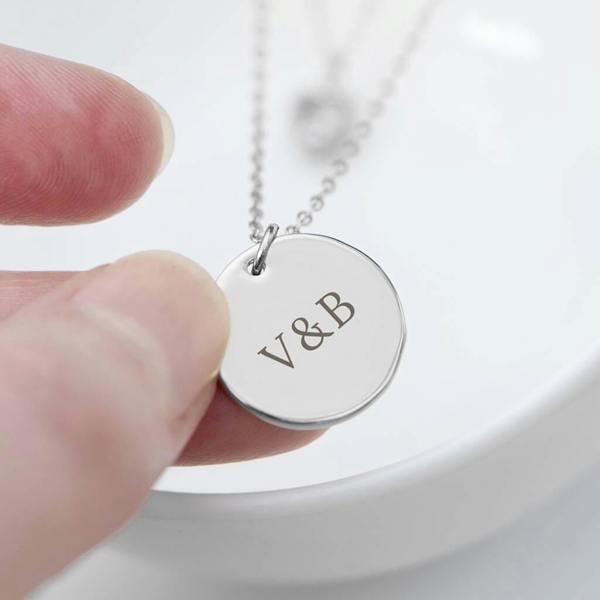 A close-up of a personalised layered silver necklace featuring a circular pendant engraved with the initials V&B held by a hand over a white bowl.