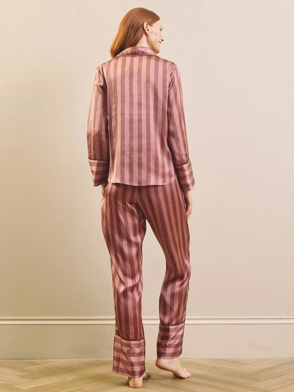 A woman stands barefoot indoors wearing a matching two-piece pyjama set with vertical stripes in dusty rose and dark brown. The set includes a long-sleeve button-up top and coordinating pants, shown from the back against a plain beige wall and light wood floor.
