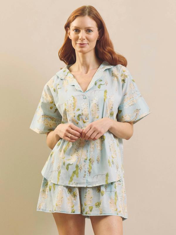 A woman modelling a light blue short-sleeve pyjama set with a green and white wisteria floral print, consisting of a collared button-up top and matching shorts, against a solid beige background.