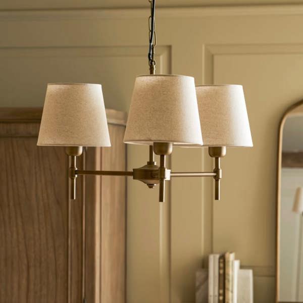 An antique bronze 3-light ceiling chandelier with cream linen shades styled in a room with an oak wardrobe and arched mirror by Abode Home & Gifts.
