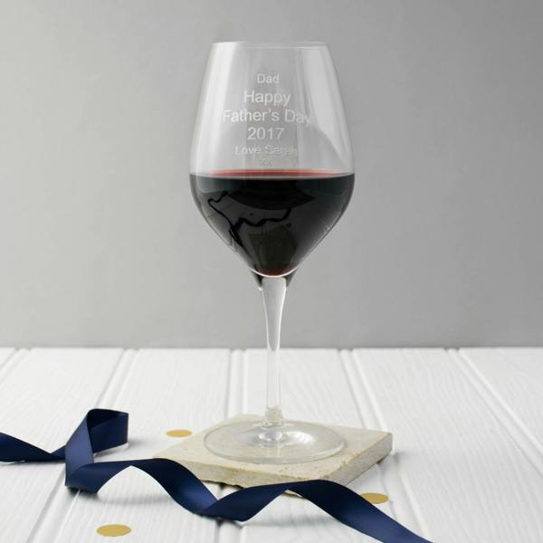 A close-up of a personalised engraved wine glass filled with red wine, resting on a stone coaster with a blue satin ribbon.