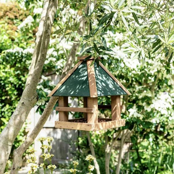 A wooden gazebo-style bird feeder with an olive green roof hanging from a leafy tree branch in a sunny garden.