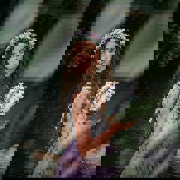 Young girl in a purple dress with a floral wreath and bouquet, standing among greenery.