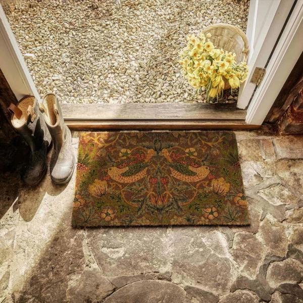 Decorative doormat with a floral pattern on a stone floor, with shoes and a basket of flowers nearby.