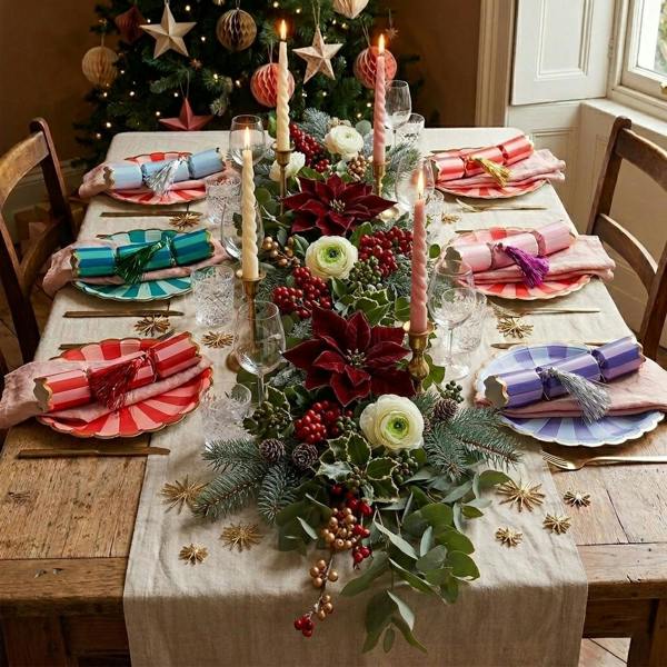 A festive dining table set for six featuring a colourful array of striped Christmas crackers with metallic tassels, deep red poinsettias, and holly foliage.