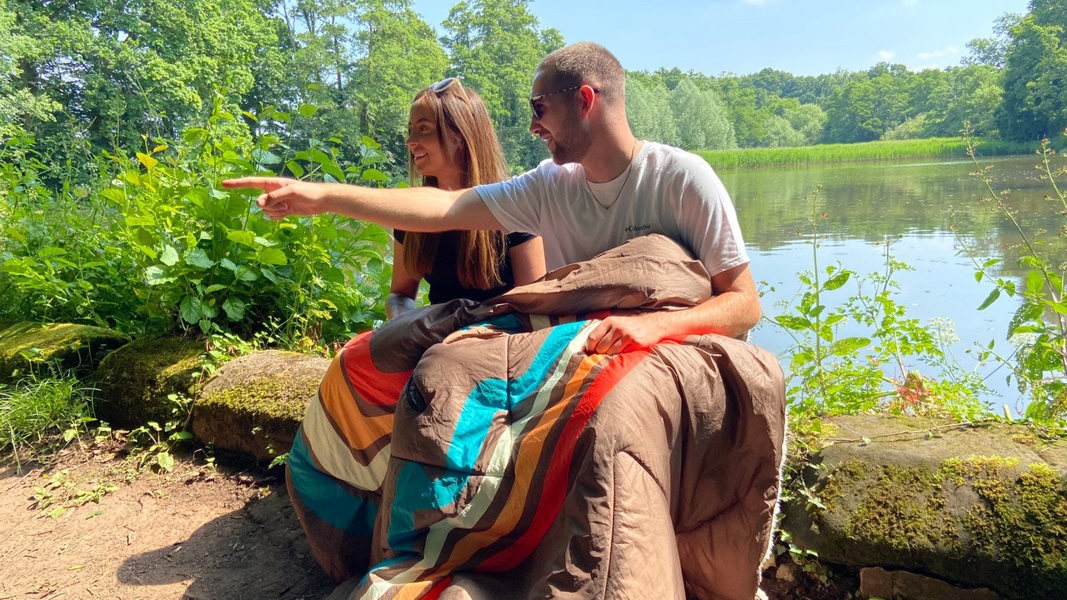 Tonal treetops and vivid spruce Insulated Outdoor Blanko been worn by a young couple enjoying the countryside sat on a log with a lake behind them, the outdoor blanket wrapped around them keeping them warm and cosy. imageset:Vivid Spruce