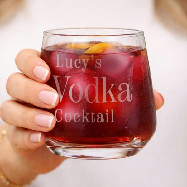 A hand holding a personalised glass tumbler filled with a dark red cocktail, ice cubes, and orange slices, showing the engraving Lucy's Vodka Cocktail.