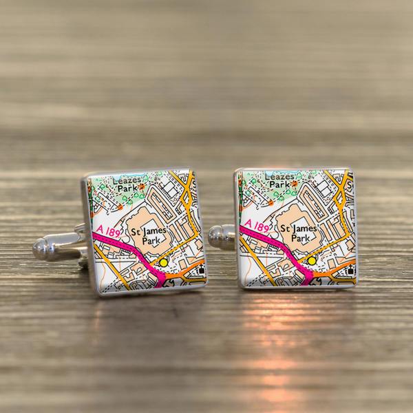 A pair of polished square silver cufflinks showing a detailed map of St James' Park football stadium on a wooden surface.