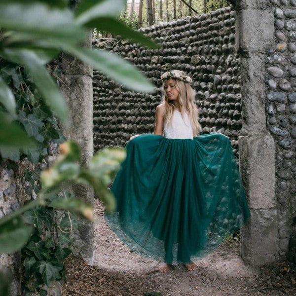 Girl in a long teal skirt standing in front of a stone wall with greenery.