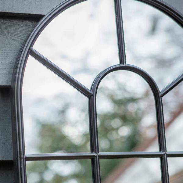 Close-up of a black metal arched window pane mirror mounted on grey exterior wood panels reflecting trees and the sky.