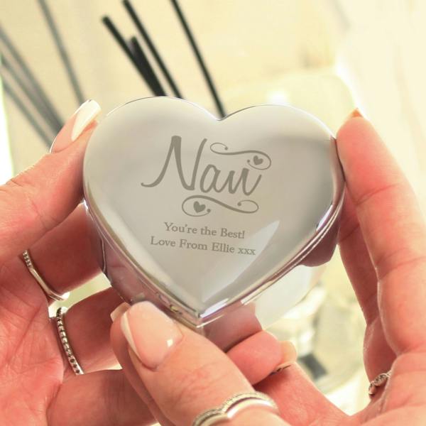 A person's hands holding a polished silver heart-shaped trinket box by Abode Home & Gifts, engraved with a decorative "Nan" swirl design and the message "You're the Best! Love From Ellie xxx" on the lid, against a soft focus background.