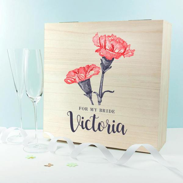 A square natural pine box printed with 'For My Bride Victoria' and pink carnations, styled upright next to two empty crystal champagne glasses and a white satin ribbon.
