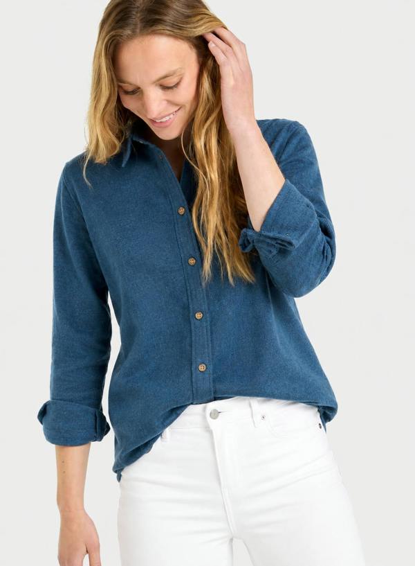 A woman wearing a blue long-sleeve button-up shirt with rolled cuffs and white pants, photographed against a light gray studio background.