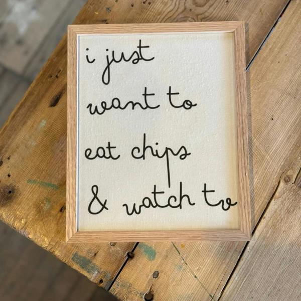 A solid oak framed white cotton typography print displaying the lowercase handwritten-style quote "i just want to eat chips & watch tv" on a rustic wooden table.