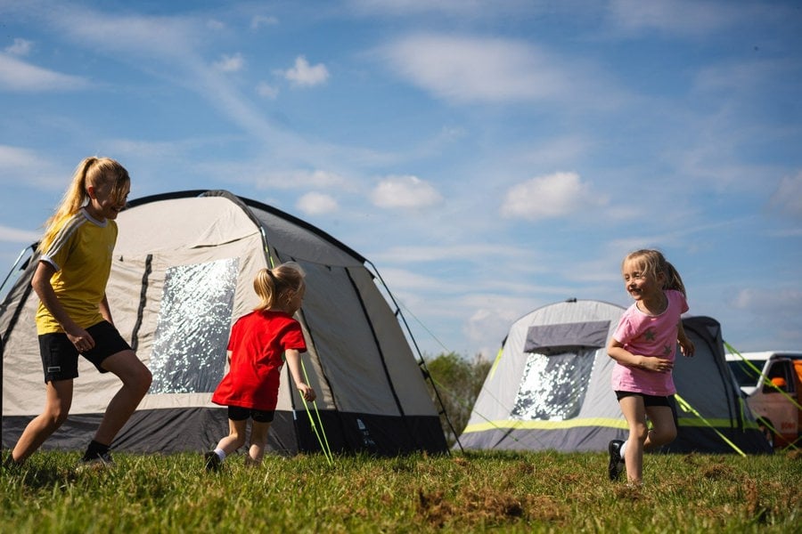 Two twin girls play with their older sister outside of the OLPRO Cubo Poled Campervan Awning on a busy campsite