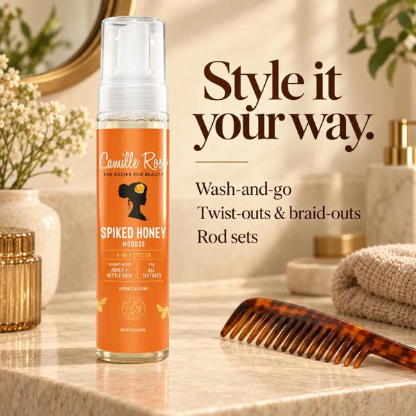 A bottle of Camille Rose Spiked Honey Mousse hair styling product stands on a marble bathroom counter beside a tortoiseshell comb, with towels, flowers, and gold-toned bathroom accessories in the background. Text on the right reads, 'Style it your way.