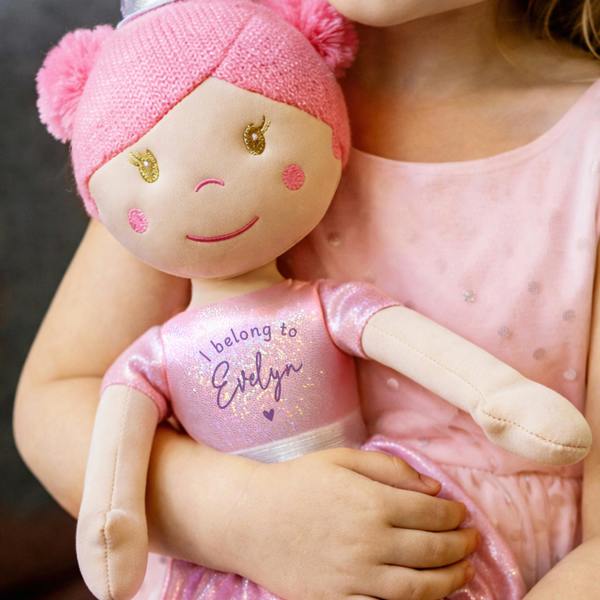 A close-up shot of a young girl in a pink sequined dress holding a plush princess rag doll. The doll has pink yarn hair, an embroidered smiling face, and wears a sparkling pink ballerina outfit text-printed with 'I belong to Evelyn' and a small purple heart.