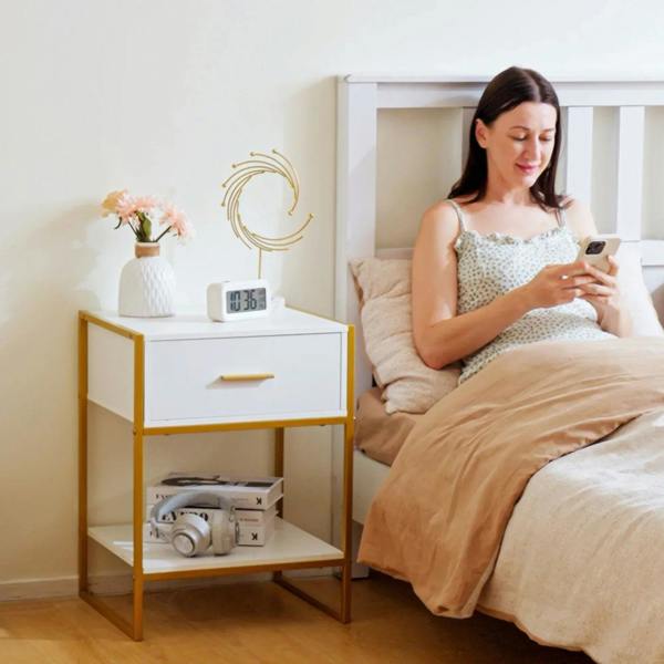 White bedside table with gold frame and drawer, open shelf holding decor and gadgets.