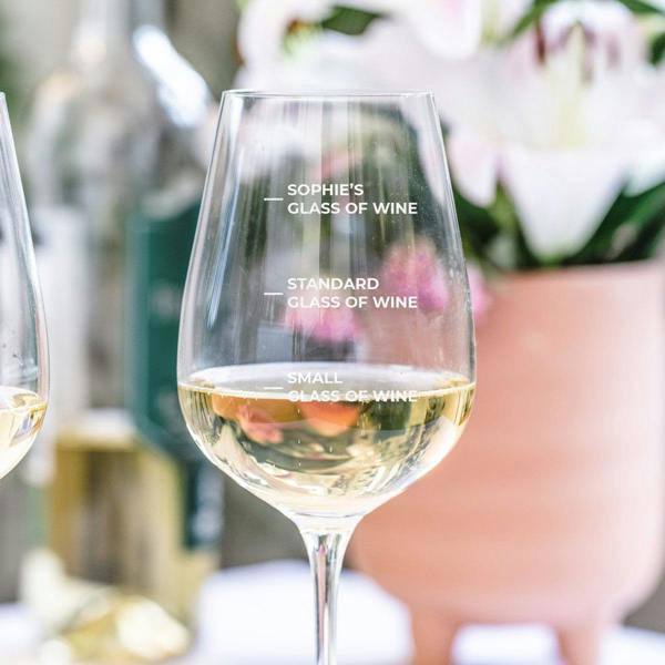 A close-up of a personalised white wine glass filled with white wine, showing three etched volume markings: Small, Standard, and Sophie's Glass of Wine at the top.
