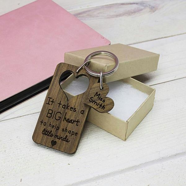 A rectangular walnut wood keyring featuring a heart-shaped cutout and the engraved words "It takes a BIG heart to help shape little minds". A smaller walnut heart charm attached to the ring is engraved with "Miss Smith". The keyring is placed on a light wooden surface next to a pink notebook and a small gift box.